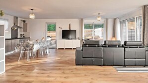 Un salon moderne doté d’un grand canapé d’angle gris, d’un coin repas avec une table et des chaises en verre, et d’une cuisine avec des meubles en bois et une cuisinière encastrée.
