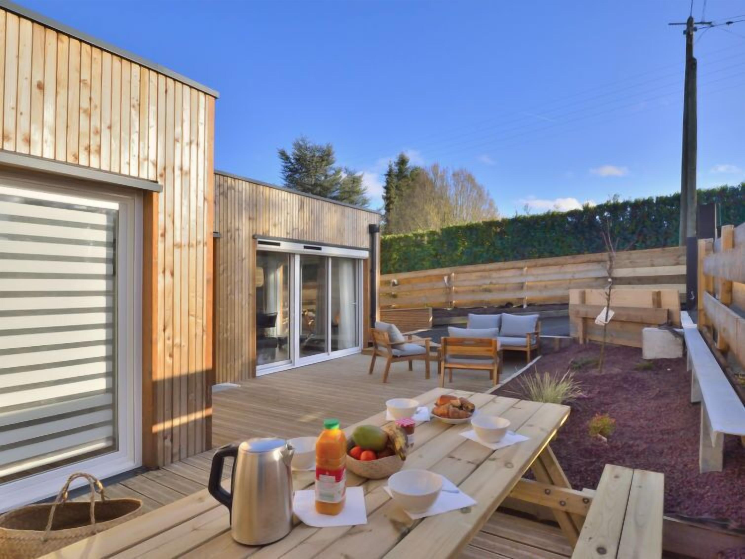 Una terraza de madera con una mesa dispuesta para el desayuno, asientos al aire libre y un gran ventanal corredizo que da a un porche cubierto.