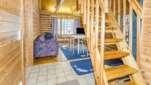 Interior de una cabaña de troncos con escalera de madera, sofá, mesa de comedor y televisor.