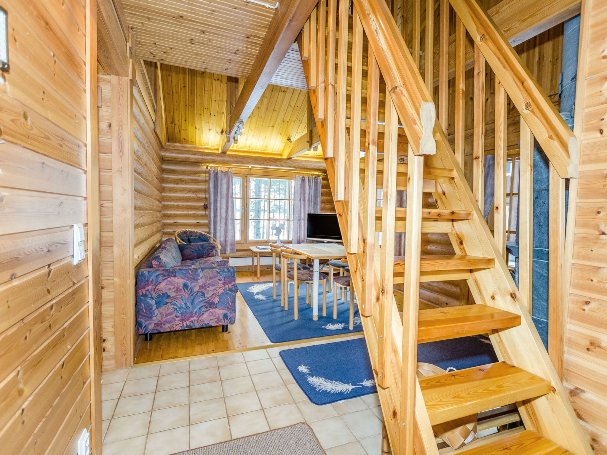 Ein Holzblockhaus-Interieur mit Treppe, einer Couch, einem Esstisch und einem Fernseher.