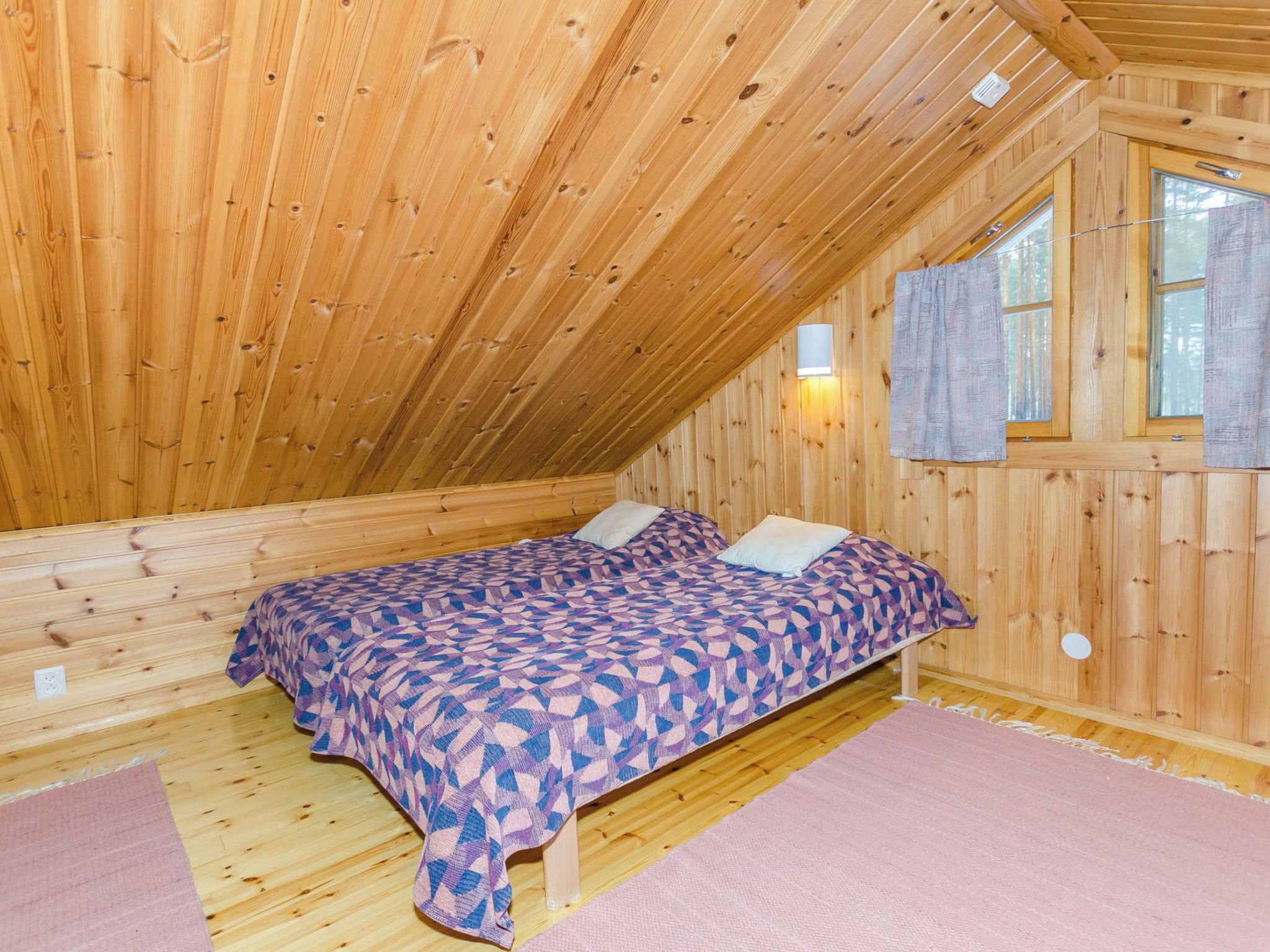 A wooden cabin room with a bed, a window, and a wall-mounted light fixture.