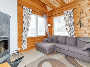 En mysig stuga med en öppen spis, en grå hörnsoffa, träväggar och mönstrade gardiner.
