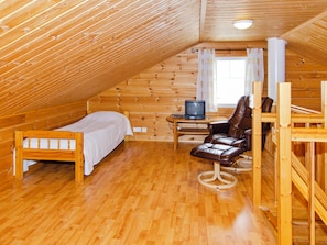 A small, cozy attic room with wooden walls and a sloped ceiling. There is a bed, a chair, and a small table with a television.