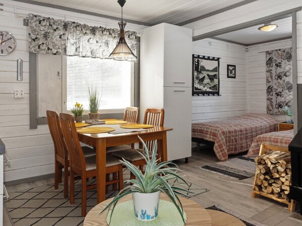 Une pièce chaleureuse avec une table à manger en bois, des chaises, un lit recouvert d’une couverture à carreaux, une horloge murale et un tableau encadré.