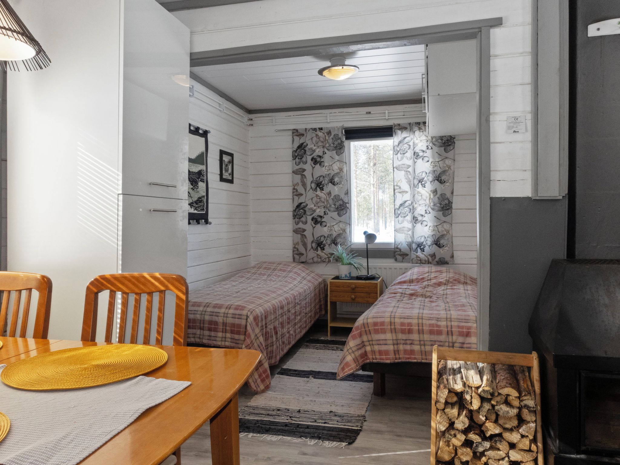 A compact room with two beds, a wooden table with a yellow placemat, a small bedside table, a radiator, and a fireplace.