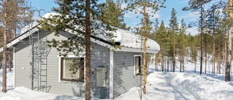 Eine kleine Holzhütte mit Satteldach, umgeben von verschneitem Gelände und hohen Kiefern.