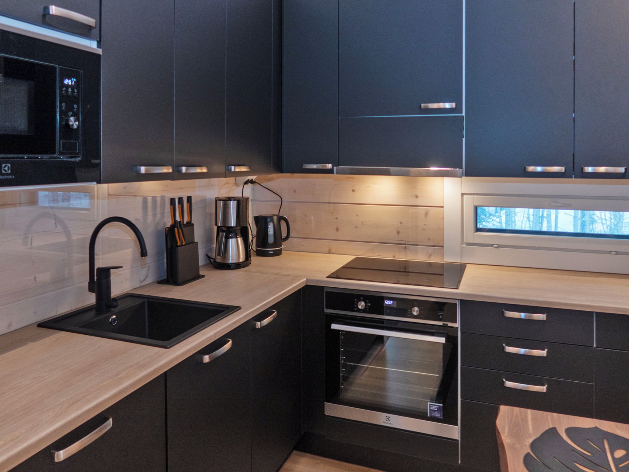 Una cocina moderna con armarios oscuros, un lavamanos de acero inoxidable, una cafetera y un horno.