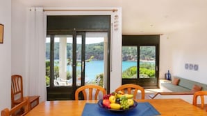 A dining area with a wooden table and chairs, a fruit bowl, and a view of a coastal landscape through large windows.