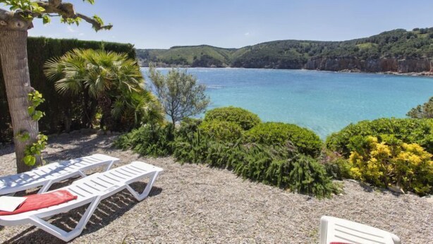 A coastal view with two white lounge chairs, a tree, and a clear blue sea.