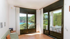 A spacious room with a checkered sofa, a wooden table, and a view of a coastal landscape through large sliding glass doors. - Port del Rei I by Interhome (L'Escala)