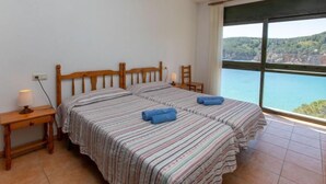 A bedroom with two beds, a nightstand, a chair, and a large window with a view of a coastline. - Port del Rei I by Interhome (L'Escala)
