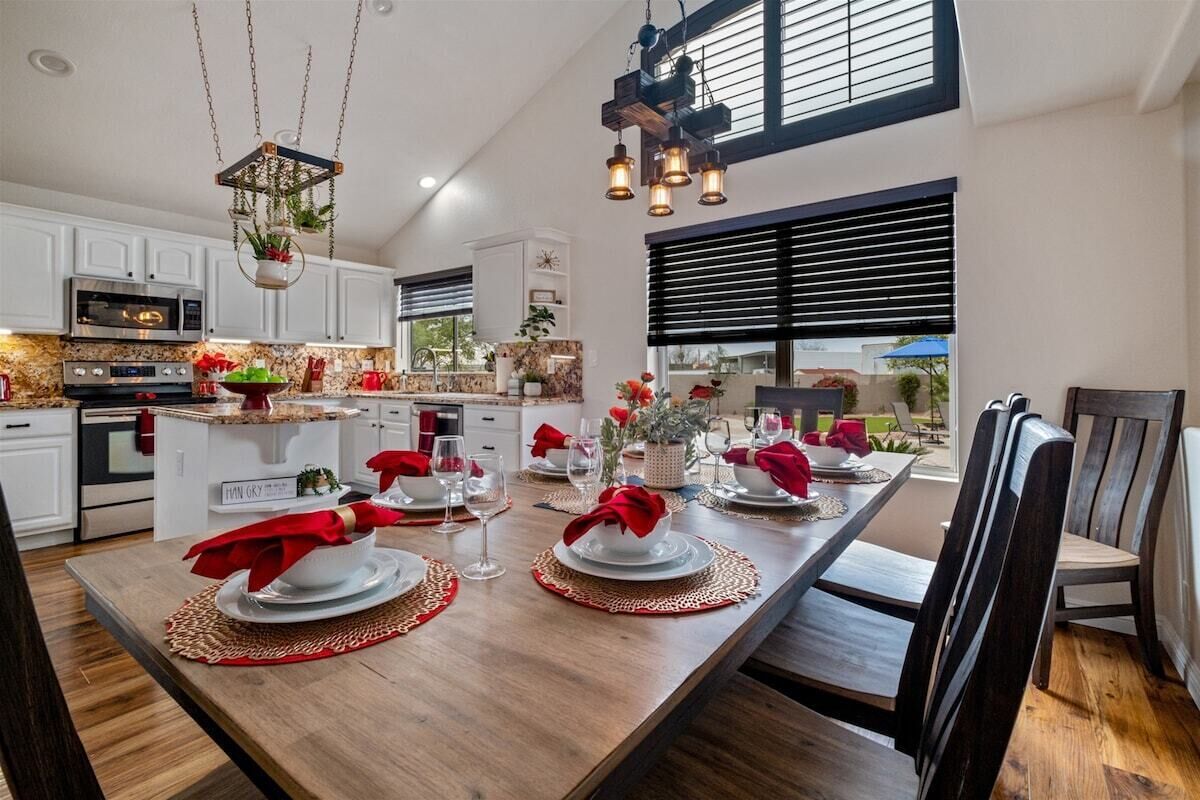 A modern kitchen with white cabinets, a built-in oven, and a dining area set for a meal with a long wooden table, red napkins, and a hanging light fixture.