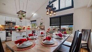 Een moderne keuken met witte kastjes, een ingebouwde oven, en een eethoek gedekt voor een maaltijd met een lange houten tafel, rode servetten en een hangende lamp.