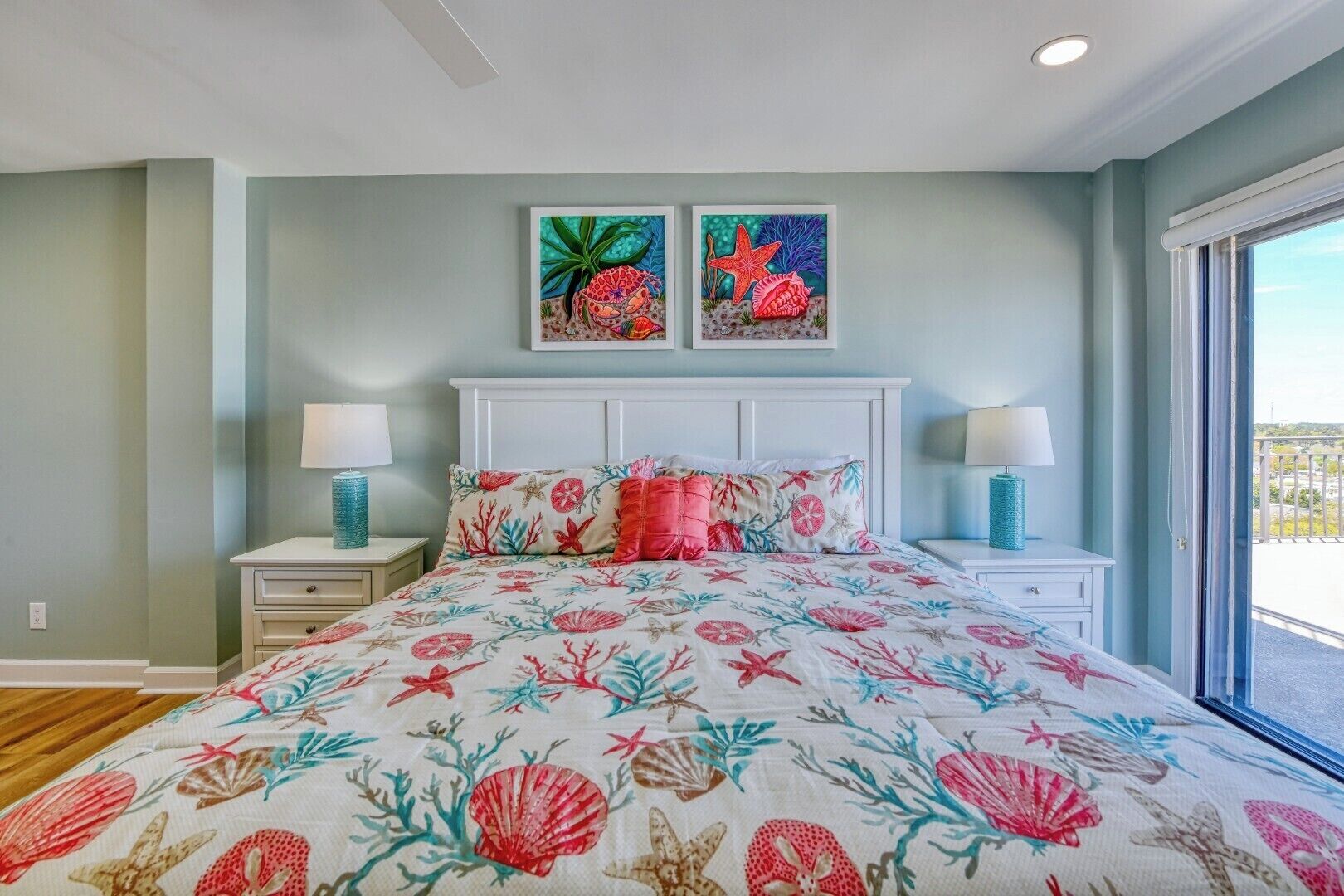 A bedroom with a floral bedspread, two wall-mounted paintings, a white headboard, bedside lamps, and a view through a window.