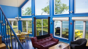 A room with a red leather sofa, a wooden table, and a yellow dining table with chairs. There are large windows and a view of the outdoors.