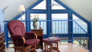 A room with a large bay window, a rocking chair, a small table with a book, and a lamp.
