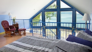 A bedroom with a large bed, a chair, a small table, and a view of the sea through a balcony.