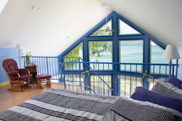 Un dormitorio con una cama grande, una silla, una mesita y vista al mar desde un balcón.