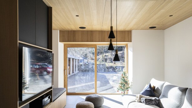 A modern living room with a large flat-screen TV, wooden ceiling, and a view of a snowy landscape through glass doors.