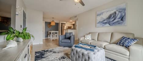 A modern living room with a beige sofa, a blue armchair, a tufted ottoman, and a large abstract painting on the wall.