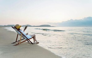 A beach with a single lounge chair and a person reading a book.
