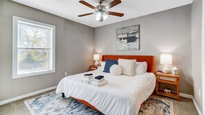A hotel room with a bed, a ceiling fan, a window with blinds, a painting on the wall, and a nightstand with a lamp. - Walkable to Oakley Square-Newly Renovated-King Bed (Cincinnati)