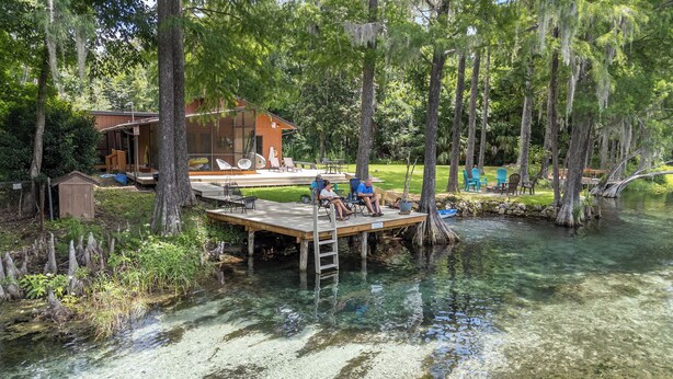 A wooden dock extending into a clear water body, surrounded by trees and a small building with outdoor seating. - Beautiful secluded setting on the Rainbow River surrounded by state forest (Dunnellon)