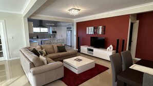 Una sala de estar moderna con un sofá seccional, una mesa de comedor con sillas, un televisor de pantalla plana y una cocina al fondo.