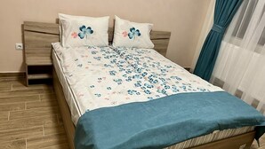 Een bed met een bloemenprintdeken, twee sierkussens met bloemenmotieven, een houten hoofdbord en een blauwe lakensprei.