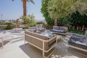 A modern outdoor seating area with sofas and a swing chair, surrounded by lush greenery and a view of distant mountains.