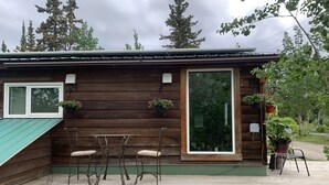 A wooden cabin with a deck, two chairs, a table, and potted plants. - Northern Flare cabin with mountain view 5 min from Nordic Eclipse (Whitehorse)