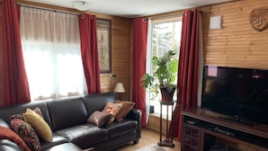 A living room with a black leather sofa, red curtains, a large flat-screen TV, a wooden cabinet, and a potted plant. - Northern Flare cabin with mountain view 5 min from Nordic Eclipse (Whitehorse)