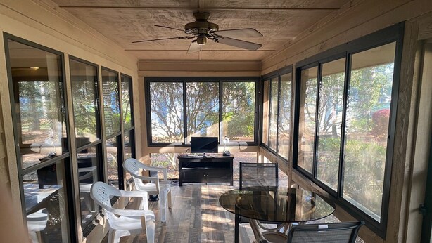 A sunlit room with a glass wall, a ceiling fan, a table with chairs, and a TV. - Dataw Island Golf Villa (Saint Helena Island)