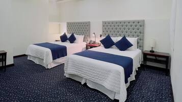 Habitación de hotel con dos camas, cada una con cabecero capitonado, ropa de cama blanca y almohadas azules decorativas. La habitación tiene una alfombra azul oscuro, una mesita de noche con lámpara y un equipo de aire acondicionado en la pared.
