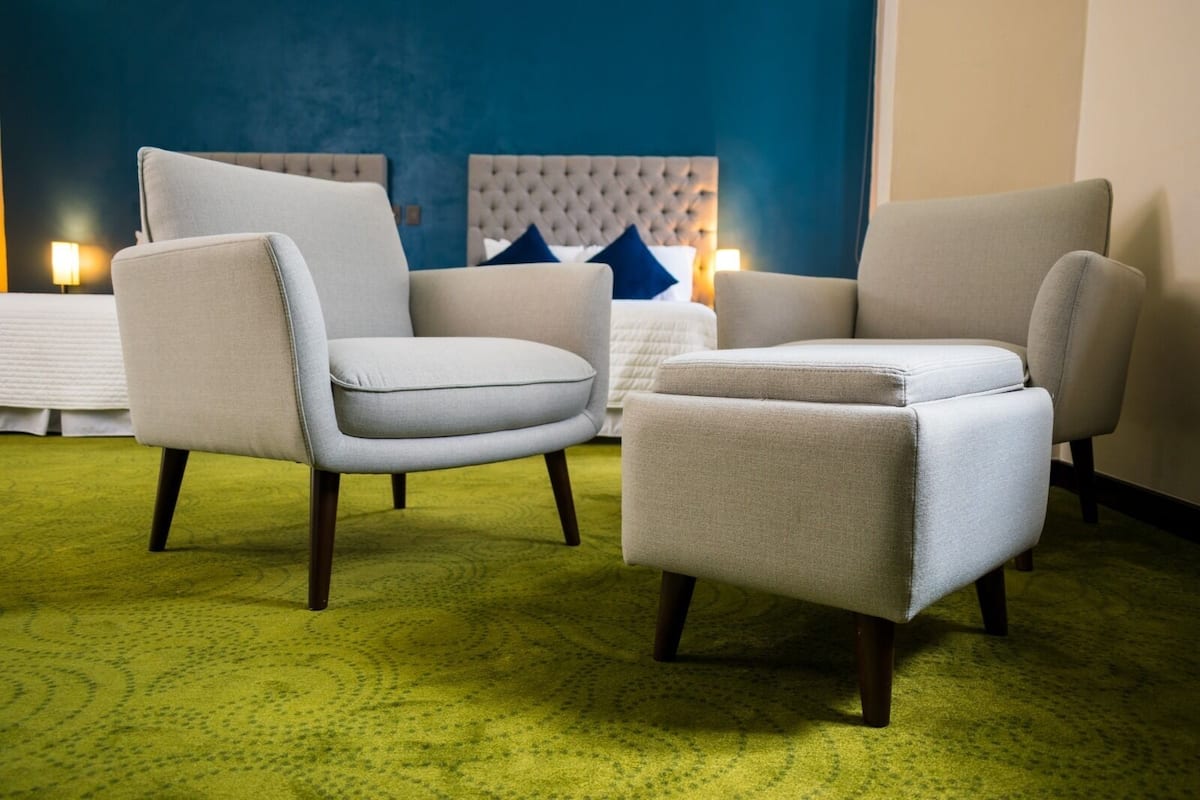 Habitación de hotel con dos sillones, un pequeño otoman, un cabecero capitonado y una alfombra verde.