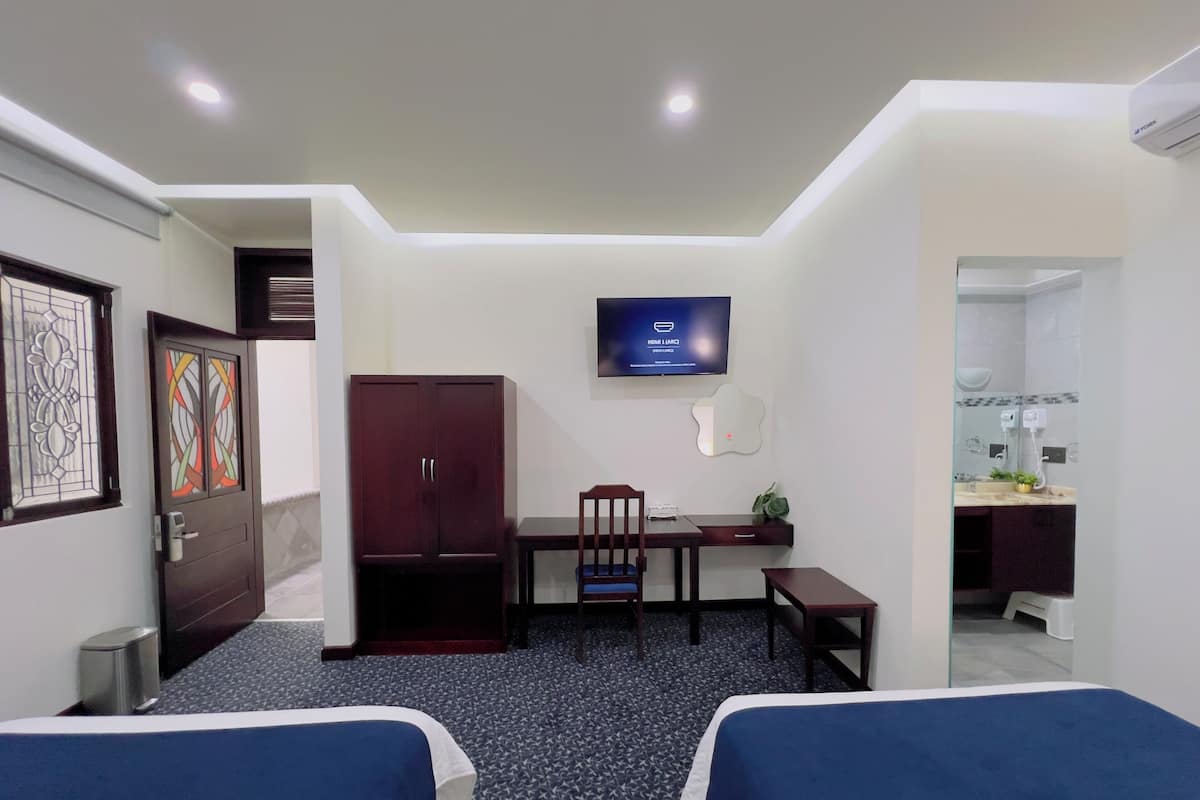 Habitación de hotel con dos camas, un escritorio, una silla, un televisor, un armario y un baño visible a través de una puerta abierta.