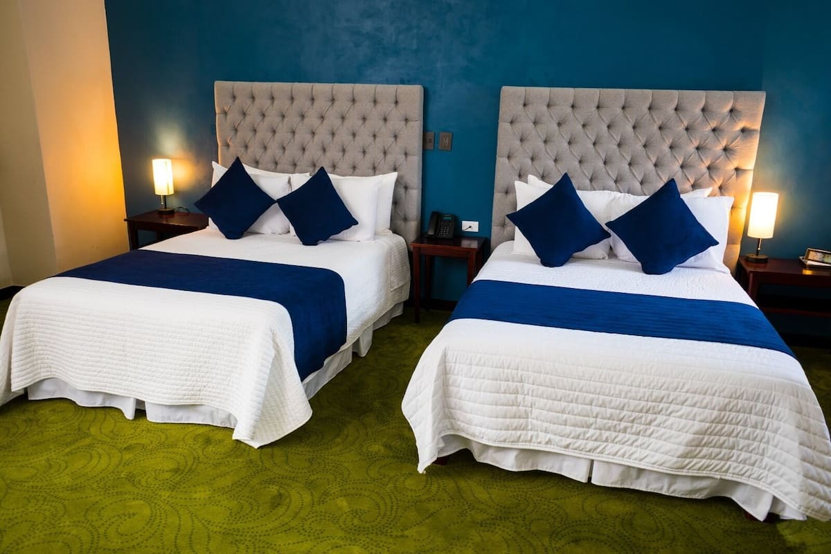 Dos camas individuales con cabeceros capitonados, ropa de cama blanca y azul, y lámparas de noche.