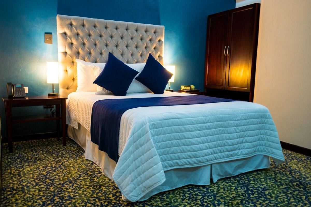 Una habitación de hotel con cabecera capitonada, cama con colcha blanca y ropa de cama azul, dos lámparas de noche, una mesita de noche y un armario de madera.