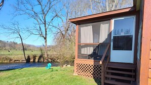 A small cabin with a wooden deck, a window with curtains, and a view of a river and trees. - Riverfront Romantic Getaway (Andrews)