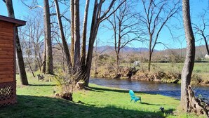 A wooden cabin with a lattice door, a grassy area with a bench, and a river in the background. - Riverfront Romantic Getaway (Andrews)