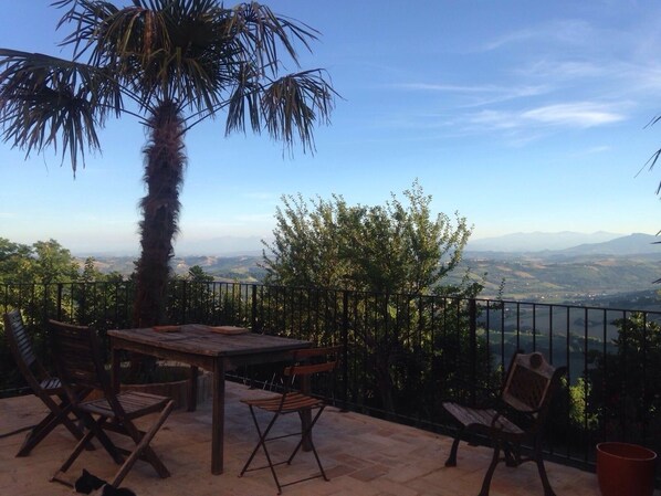 An outdoor patio with a wooden table and chairs, a palm tree, and a view of distant mountains and a valley.