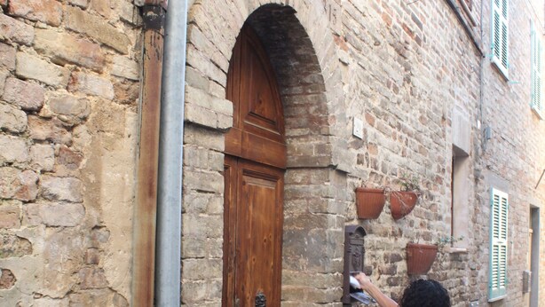 A person standing in front of a stone building with a wooden door and a small window with shutters.