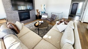 A modern living room with a large sectional sofa, a flat-screen TV mounted on the wall, a fireplace, and a dining area with a glass table. - Best View on Fairway, With ready Hot Tub! (Park City)