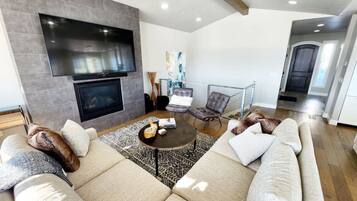 A modern living room with a large sectional sofa, a flat-screen TV mounted on the wall, a fireplace, and a dining area with a glass table.
