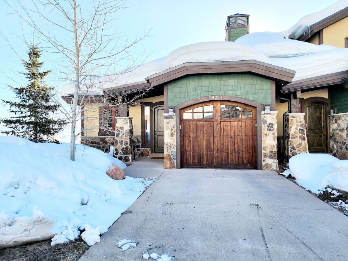 A snow-covered house with a stone facade, a wooden front door, and a chimney.