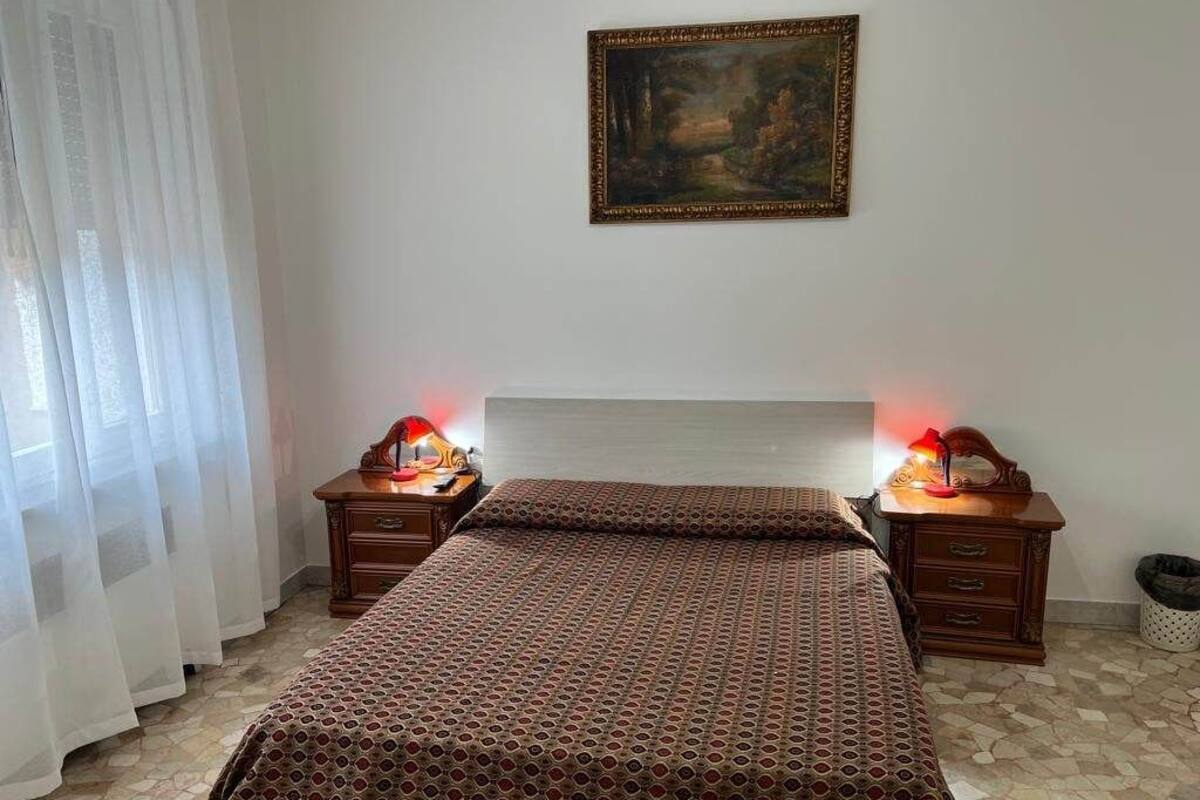 A bedroom with a patterned bedspread, two bedside tables with lamps, a painting on the wall, and a window with sheer curtains.
