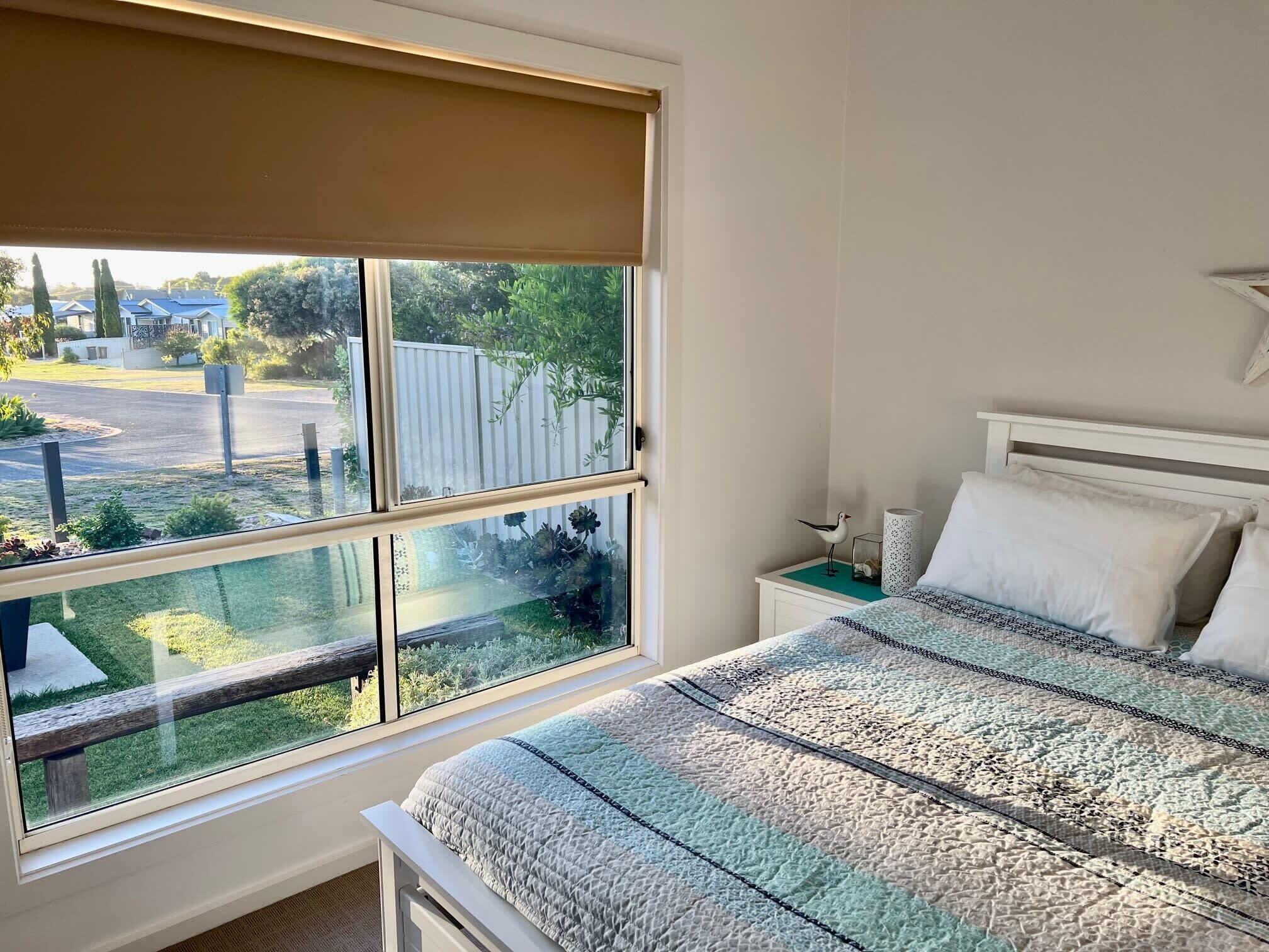 A bedroom with a large window, a bed with patterned bedding, a bedside table, and a wall-mounted lamp.
