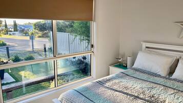 A bedroom with a large window, a bed with patterned bedding, a bedside table, and a wall-mounted lamp.