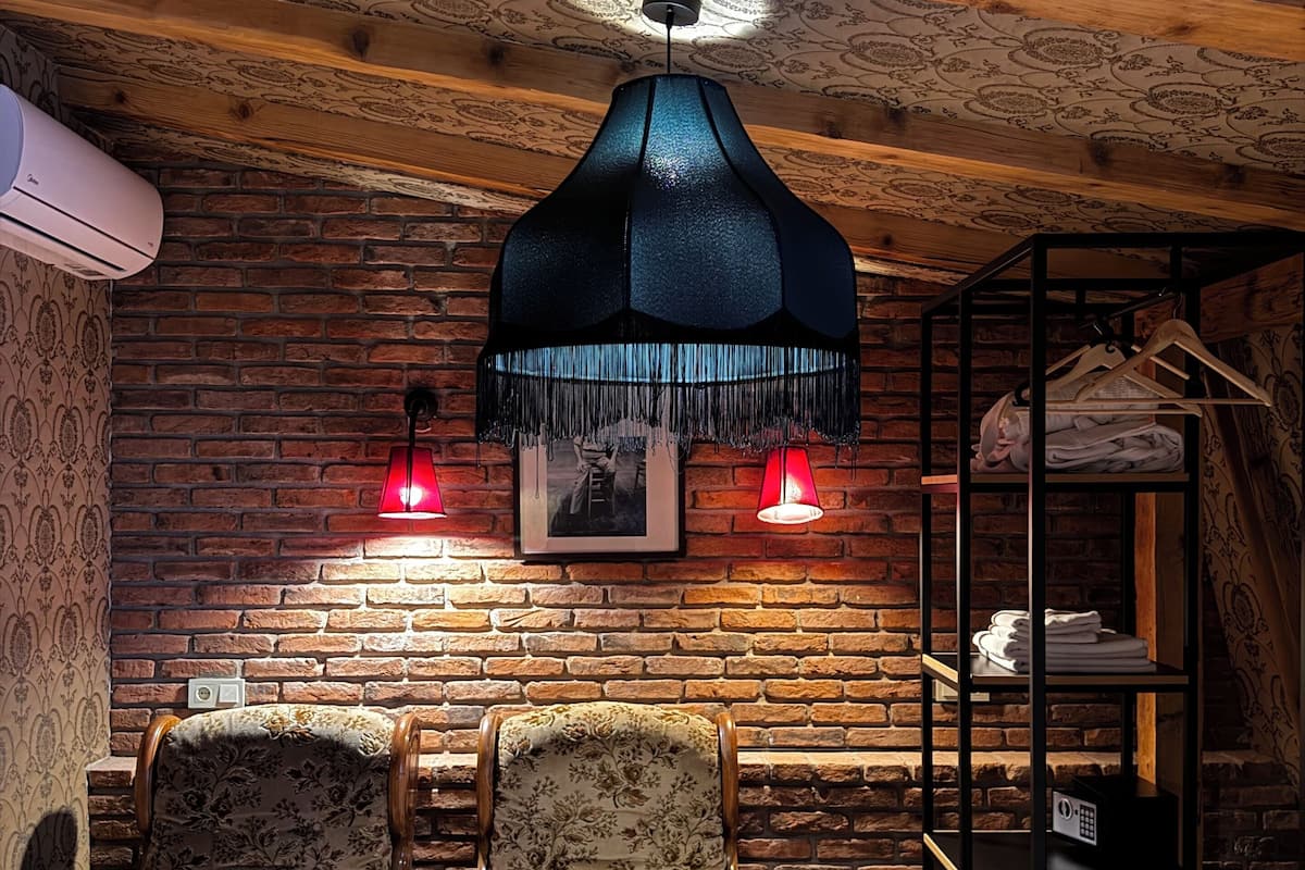 Quarto com parede de tijolos, duas poltronas estofadas, um candeeiro pendente preto e uma estante de madeira com roupas.