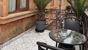 An outdoor seating area with a glass table and wicker chairs, a potted plant, and a building with large windows and stone walls. - Memoire (Kutaisi)